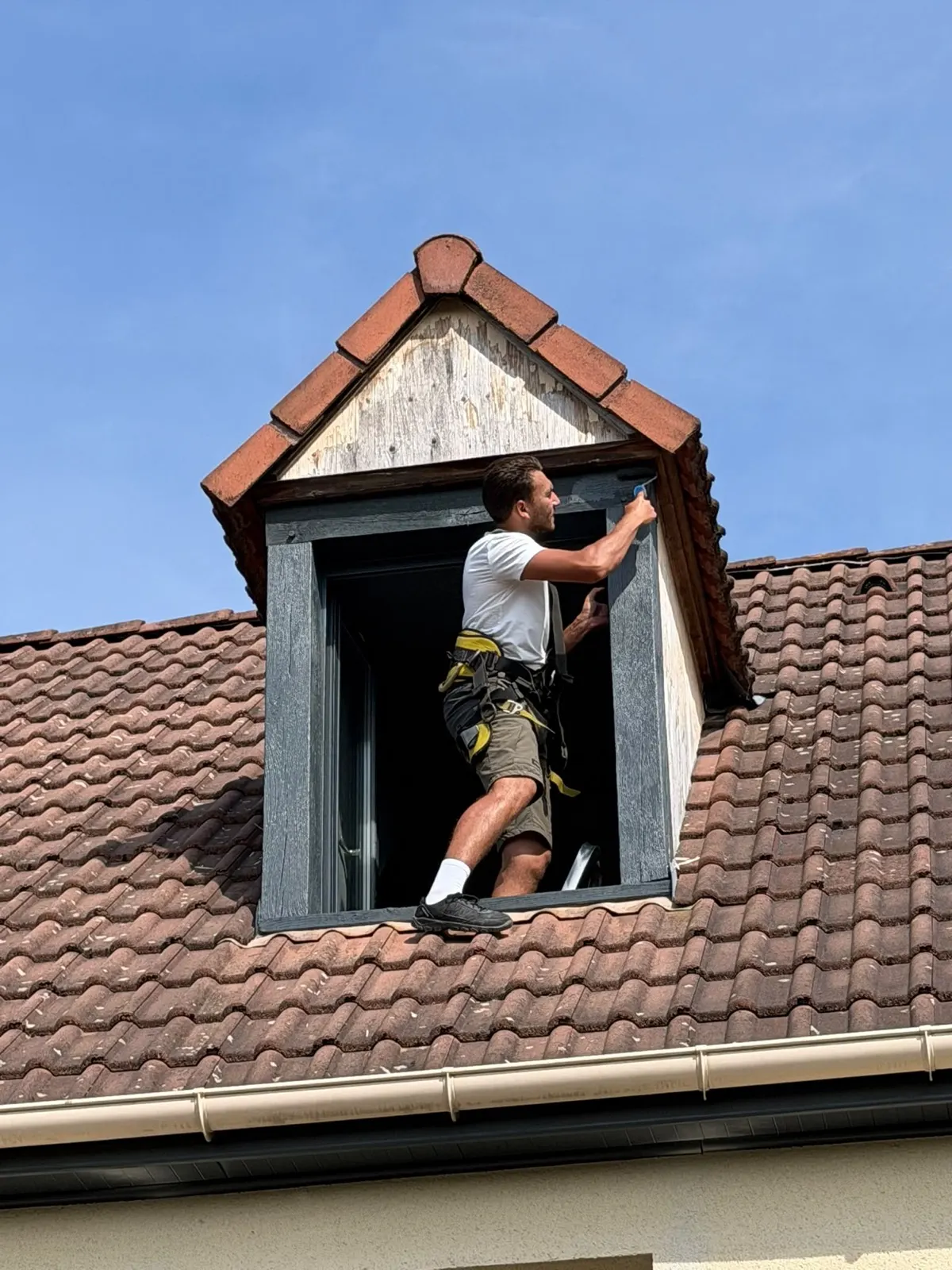 Couvreur en harnais peignant l'encadrement d'une lucarne de toit près de Rouen